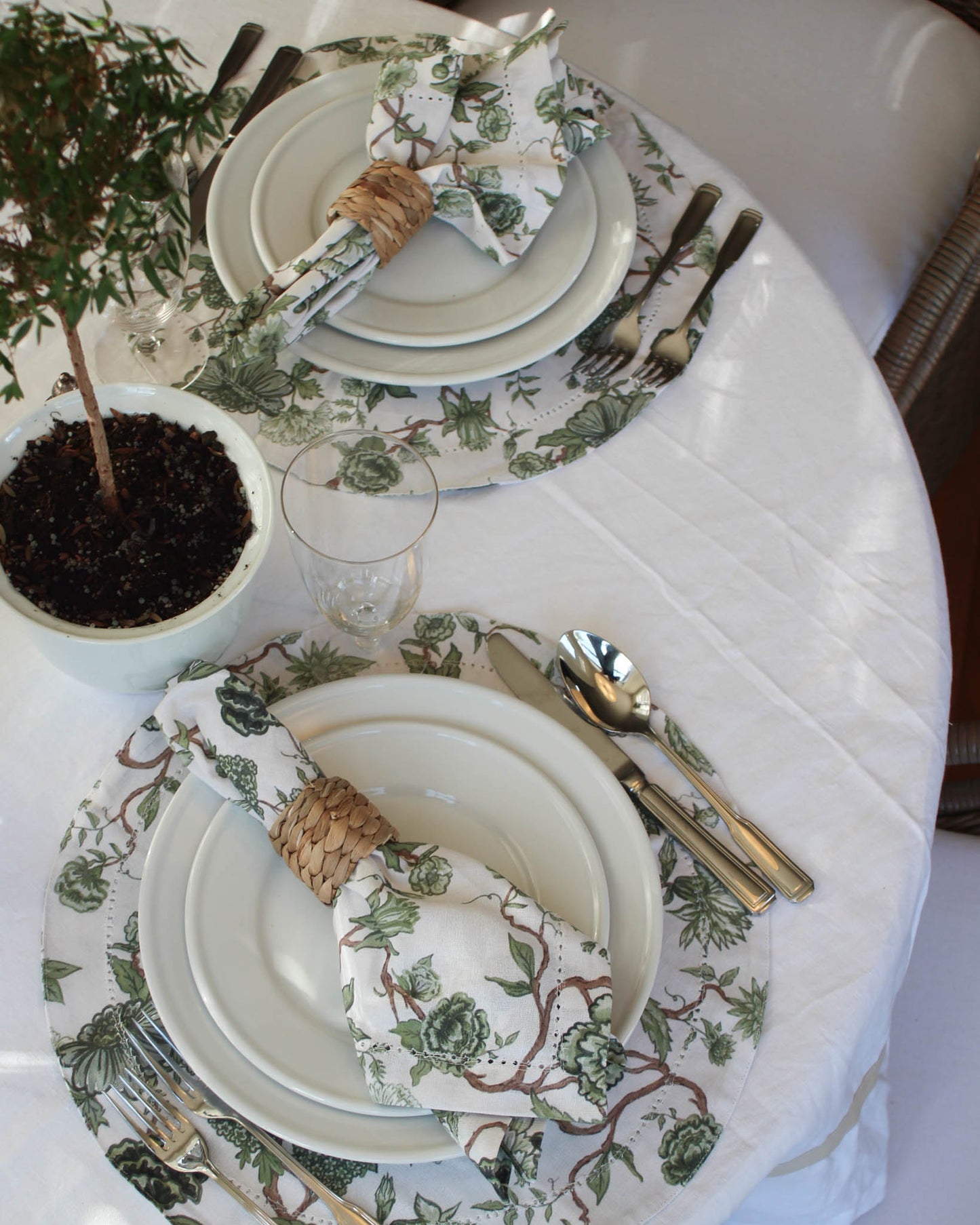 Elegant table setting with green floral napkins, cutlery, and a small plant on a white tablecloth.