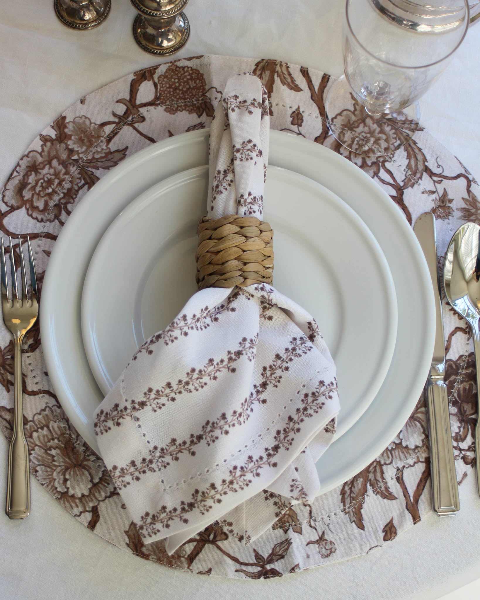 Table setting with floral napkin, white plate, and silverware on a patterned brown floral tablecloth.