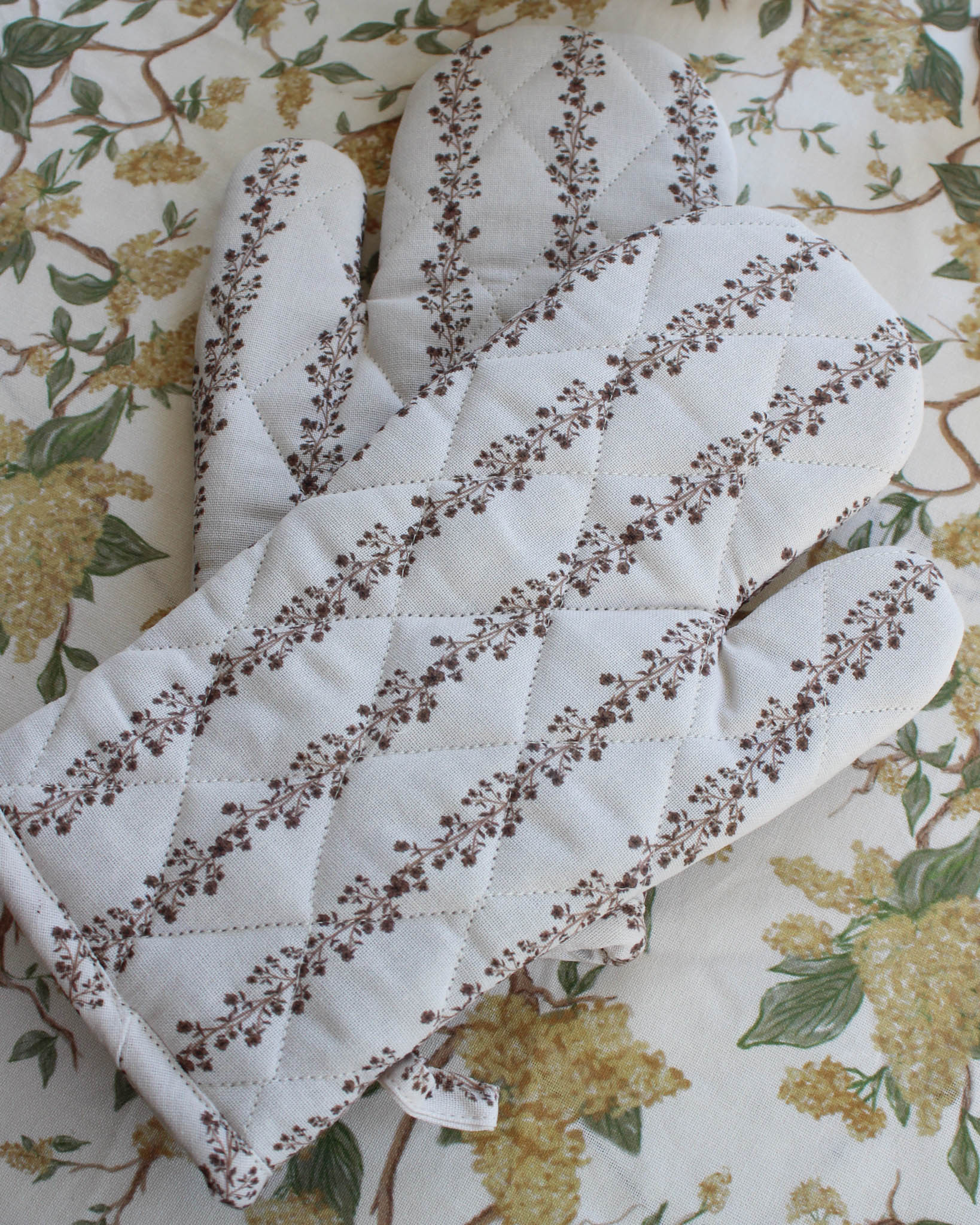 Pair of quilted oven mitts with floral print on a floral yellow tablecloth