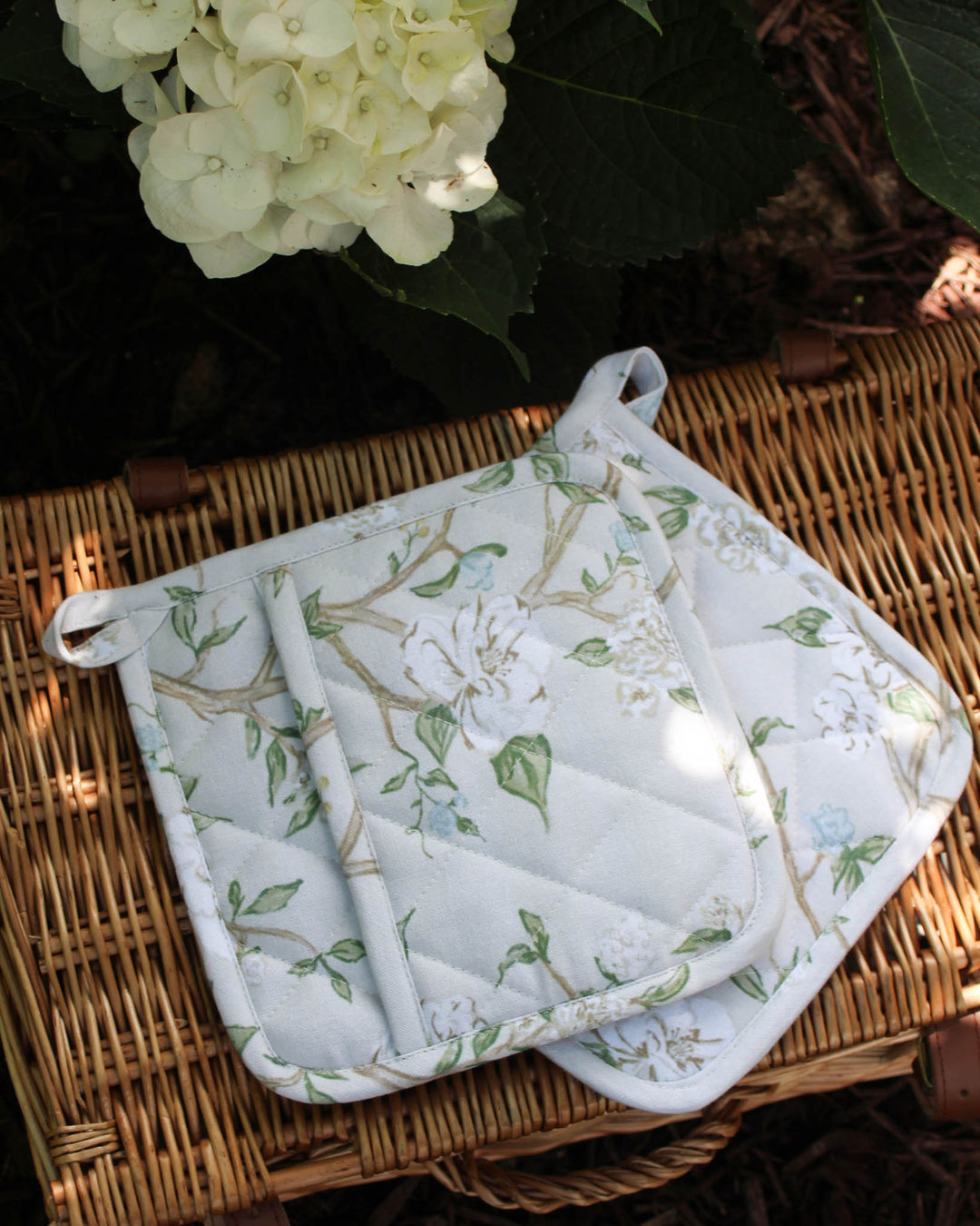 Floral-patterned pot holder and trivet on a woven surface with flowers in the background