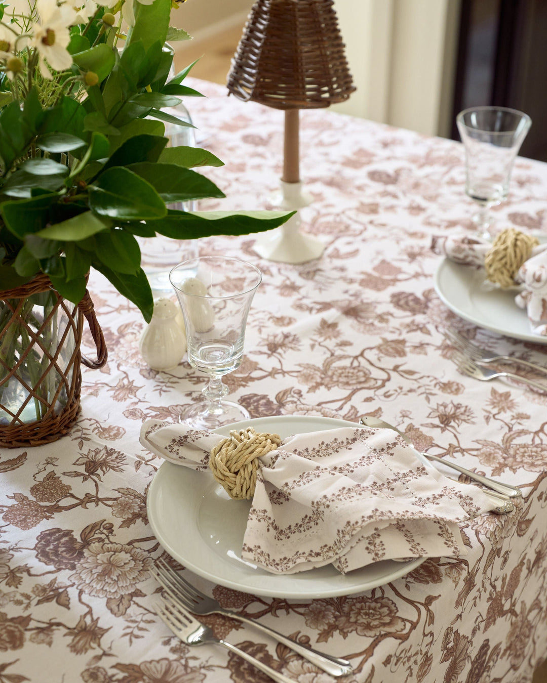 A close-up of a dining table set with a beige tablecloth featuring a brown floral toile-style pattern. The setting includes white plates, silver cutlery, a folded cream napkin with a brown floral print and a woven straw napkin ring, a glass, and a wicker basket vase with green leaves and white flowers.