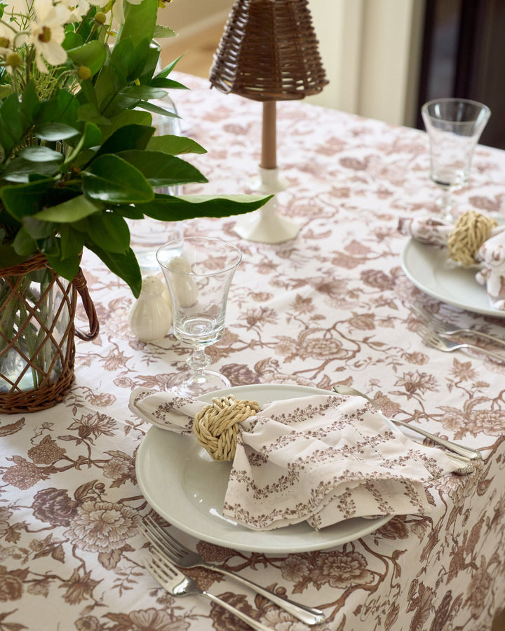 A close-up of a dining table set with a beige tablecloth featuring a brown floral toile-style pattern. The setting includes white plates, silver cutlery, a folded cream napkin with a brown floral print and a woven straw napkin ring, a glass, and a wicker basket vase with green leaves and white flowers.