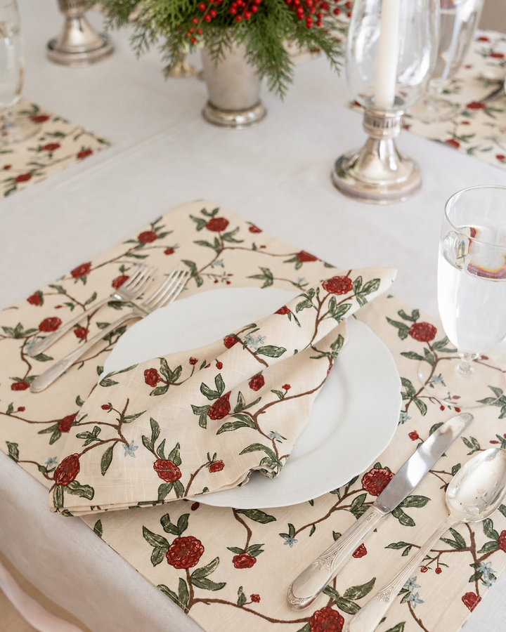 A styled table setting featuring beige linen napkins with a red berry and green leaf branch pattern, folded around white plates with silver cutlery on a white tablecloth.