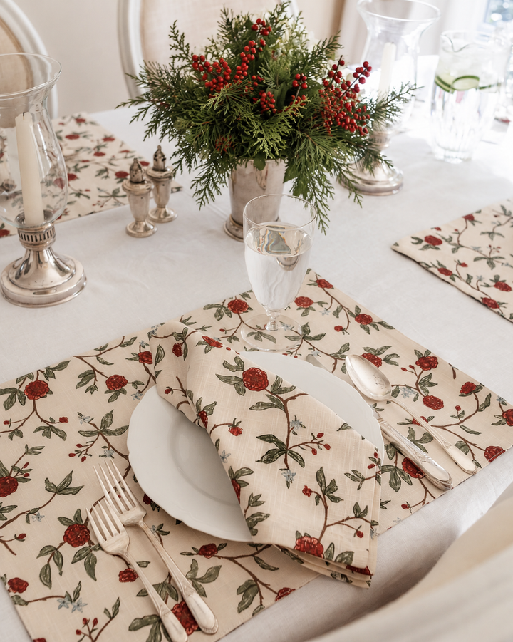 A styled table setting featuring cream table linens with a red berry and green leaf floral pattern, including a folded napkin and matching placemat, set with white plates, silver cutlery, and a central green and red floral centerpiece.