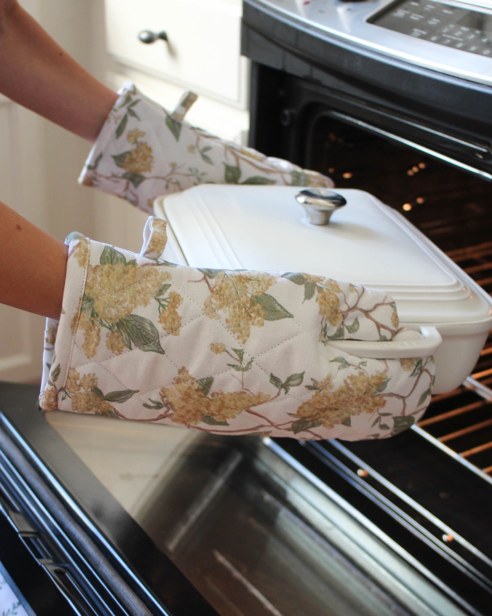 Person wearing floral oven mitts holding a covered dish in front of an open oven.