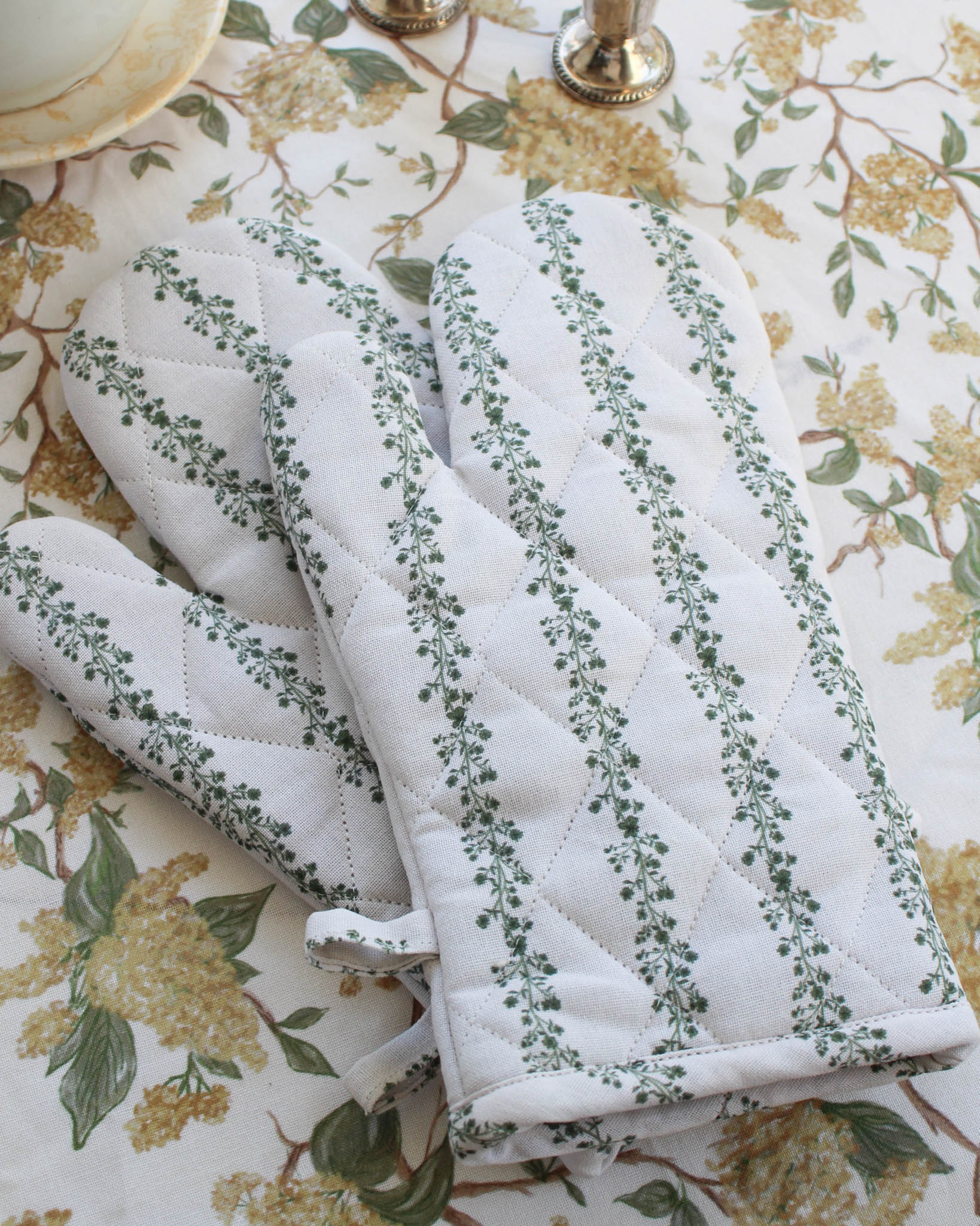 White and green floral quilted oven mitt and pot holder with green leaf pattern on a floral tablecloth.