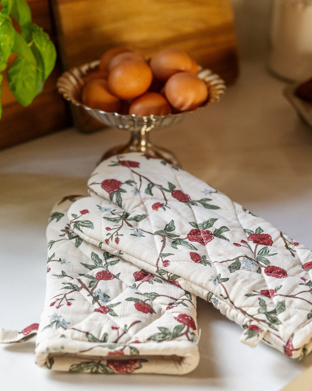 A pair of quilted red floral oven mitts that fit perfectly into a traditional farmhouse or rustic kitchen. The dense botanical pattern of green leaves and red blossoms offers a cozy, lived-in feel. Made from durable organic cotton, these long kitchen gloves are built for daily use. The recycled cotton filling ensures hands stay cool while handling hot cast iron or stoneware. A timeless accessory that blends heritage design with modern functionality.