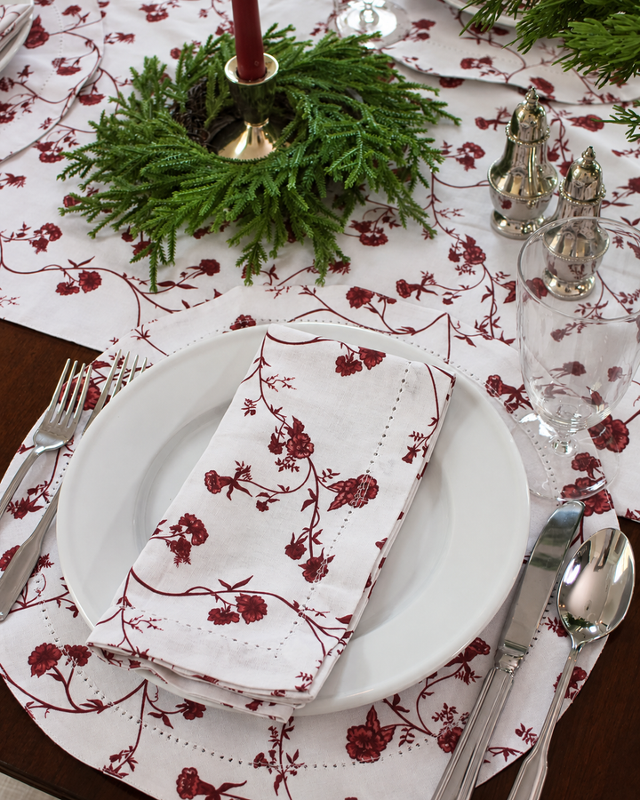 Christmas table setting featuring red floral cotton napkins and matching placemats, white dinnerware, silver flatware, and fresh evergreen greenery for traditional holiday entertaining