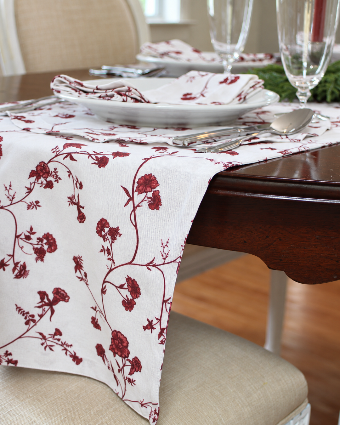 Red floral cotton Christmas table runner styled on a dark wood dining table with matching floral napkins, evergreen centerpieces, and red taper candles for festive holiday hosting.