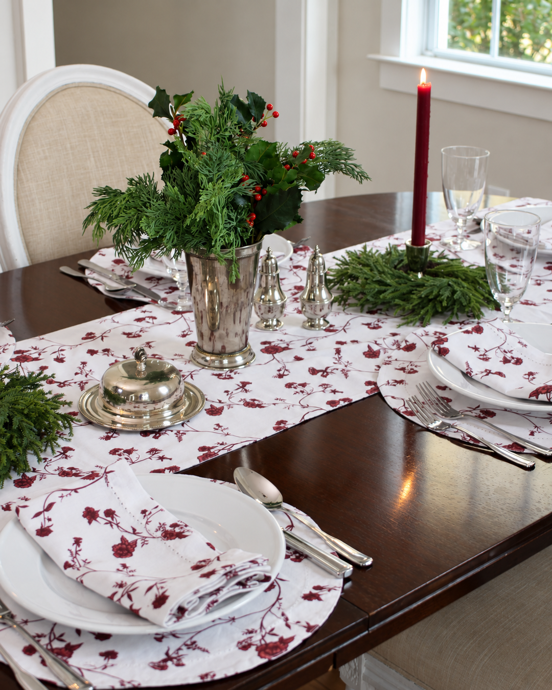Red floral cotton Christmas table runner draped over a wooden dining table, styled with matching floral napkins, white dinnerware, and silver flatware for elegant holiday hosting.