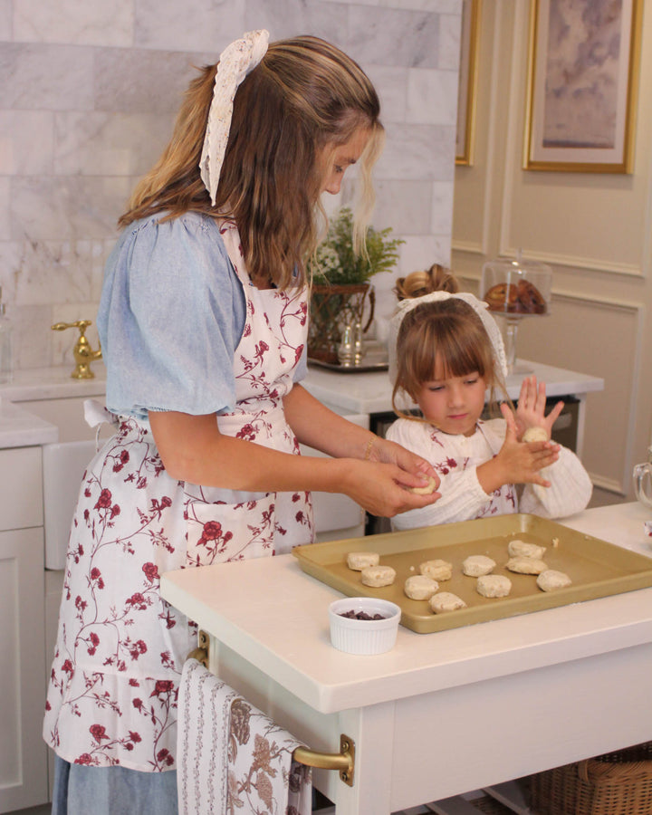 Ruffle cooking apron in a red floral print, featuring a smock apron dress silhouette. This 100% cotton apron is designed with two pockets and adjustable ties for a one-size-fits-most fit. Ideal for use as a baking apron for women, gardening apron, or BBQ apron. This red floral apron dress is perfect for spring summer entertaining and festive Easter baking. A high-quality gift for her, mom, or a baker, it adds warmth and elegance to any kitchen with its grand millennial decor style.