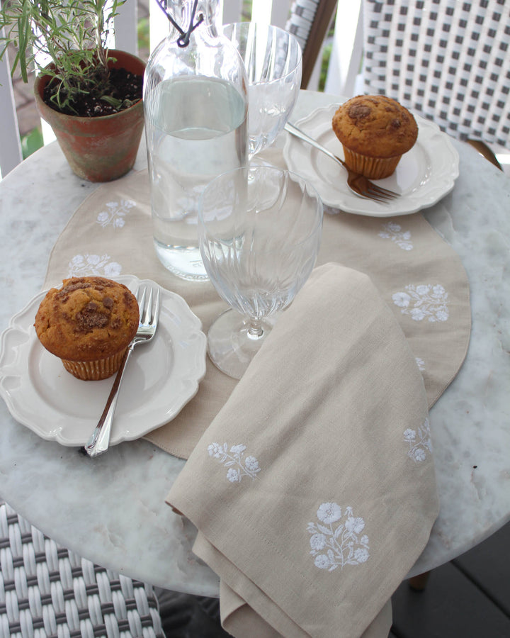 A beautiful top-down view of a beige linen-cotton round placemat featuring white floral embroidery, perfectly paired with a white scalloped plate and a rustic woven napkin ring. These placemats are designed for the grand millennial who loves a detailed, layered table for spring. Ideal for a Mother's Day brunch or garden wedding, these 15-inch mats are both functional and decorative. Give them as a gift for a hostess or use them to elevate your own country-chic Easter table decor.