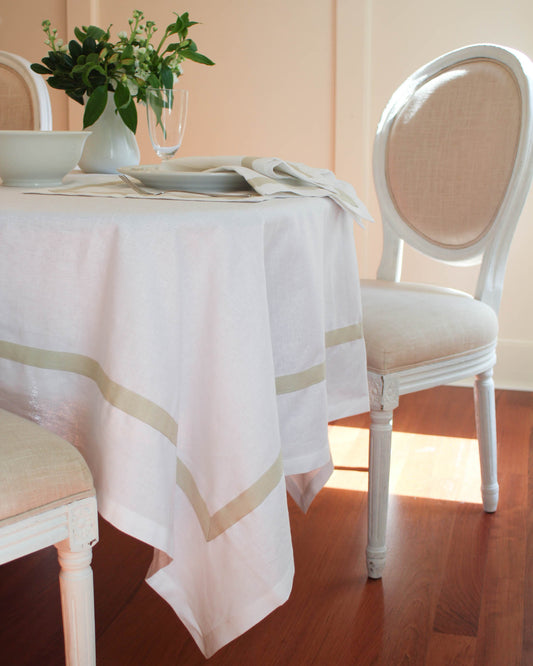 A white linen tablecloth drapes over a table, featuring an embroidered monogram  within a decorative floral border near the edge. The tablecloth has beige border that adds to elegance.