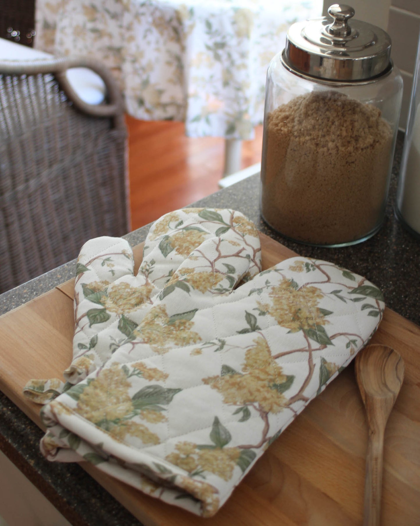 Floral-patterned oven mitt and pot holder on a wooden cutting board with a glass container of sugar in the background.