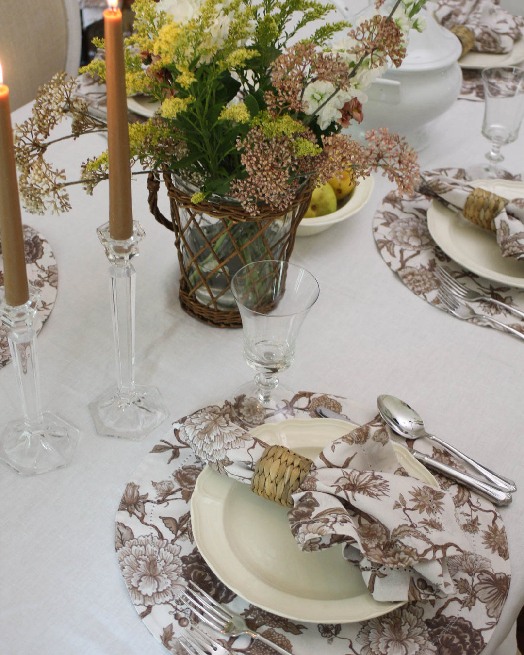 Elevate your spring table with these brown floral round placemats. Featuring a sophisticated Chinoiserie-inspired print of leafy vines and earth-toned blossoms, these cloth placemats bring a grounded, grand millennial charm to your home. The elegant hemstitched edge adds a tailored, artisan finish. Perfectly sized for spring hosting, these washable cotton placemats pair beautifully with white scalloped china and woven accents for a textured, layered French country table setting.