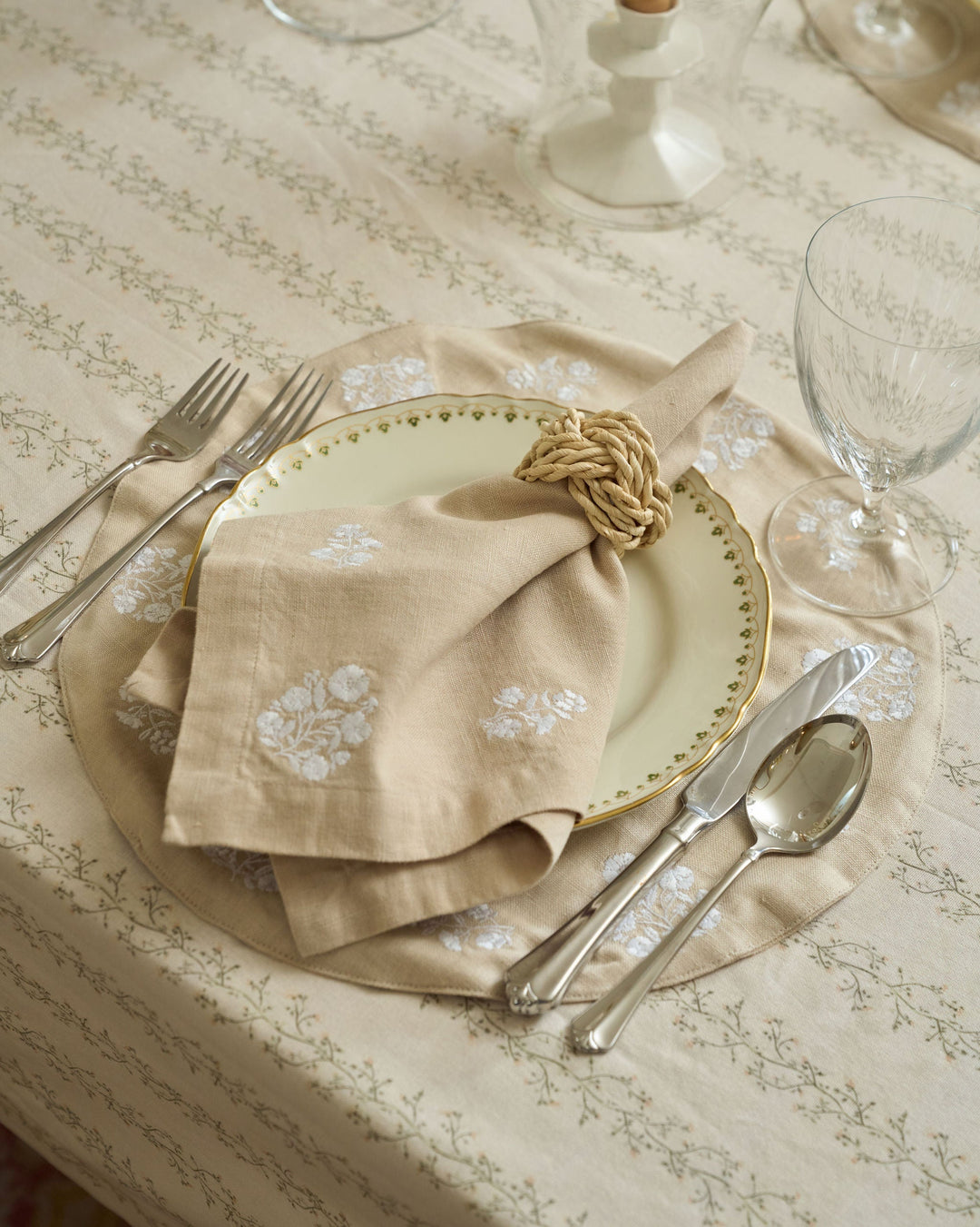 A styled table setting featuring a beige linen napkin with white floral embroidery, folded over a gold-rimmed plate, with silver cutlery and a glass on a patterned tablecloth.