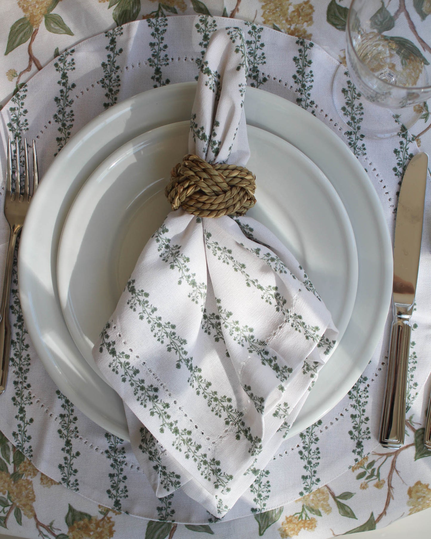 Set table with white plates, floral-patterned napkin and round placemat, and cutlery on a floral tablecloth.