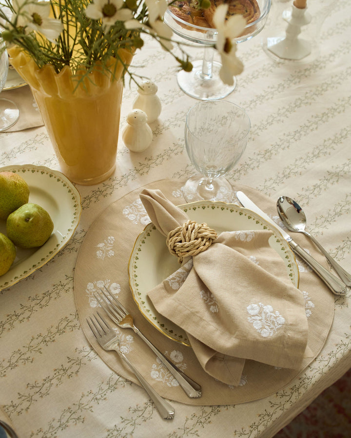 A styled table setting with a beige floral tablecloth, matching placemats, white plates with green trim, a cream napkin tied with a woven ring, silver cutlery, a yellow vase of white flowers, and a plate of green pears.
