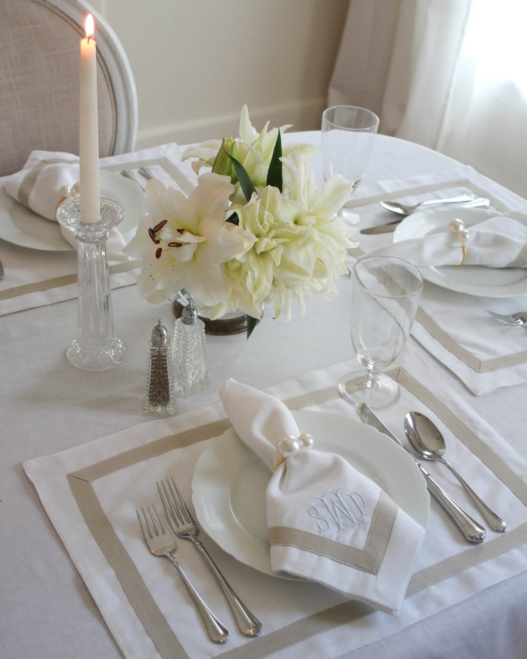 A styled dining table setting with white plates, silver cutlery, folded white napkins with monogram embroidery, a clear candlestick with a lit candle, and a floral centerpiece on a white tablecloth.
