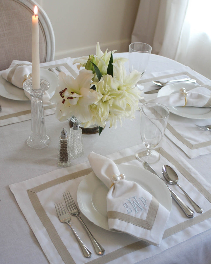 A styled dining table setting with white plates, silver cutlery, folded white napkins with monogram embroidery, a clear candlestick with a lit candle, and a floral centerpiece on a white tablecloth.