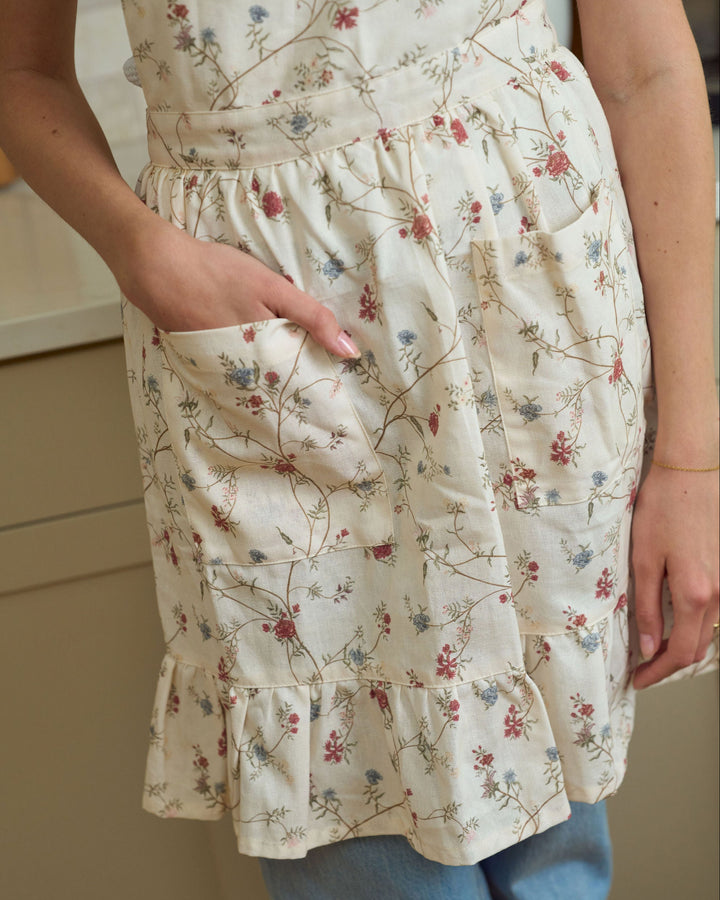 Cream floral cooking apron with feminine ruffle details, a softly gathered waist, front pockets, and adjustable ties, perfect for spring baking, summer hosting, autumn cooking, Thanksgiving gatherings, Christmas kitchens, and year-round entertaining.
